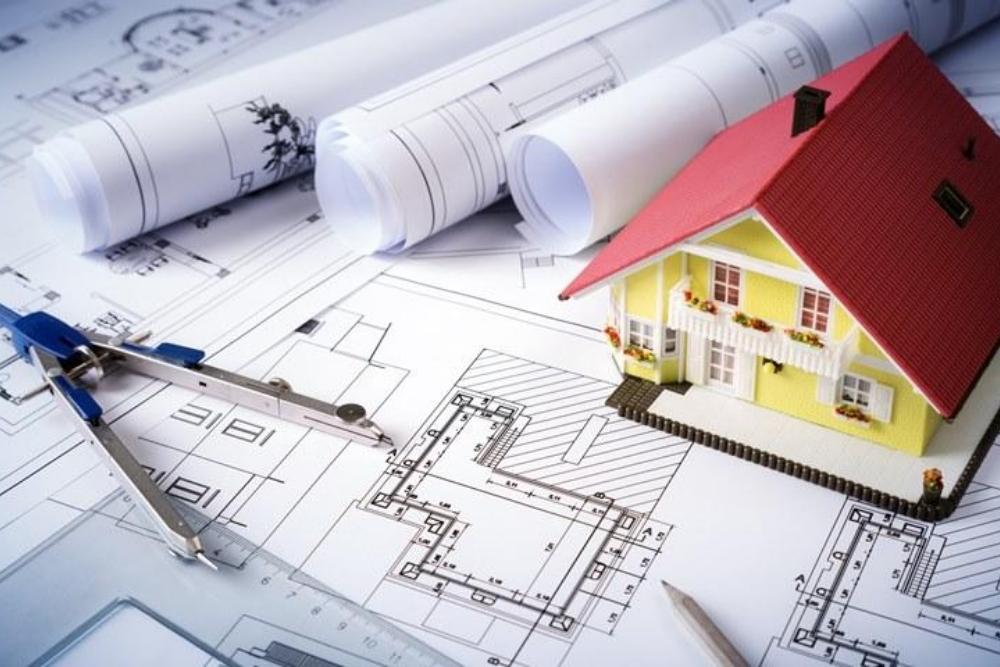 Architectural blueprints spread out with a small yellow model house, a compass, and rolled-up plan-